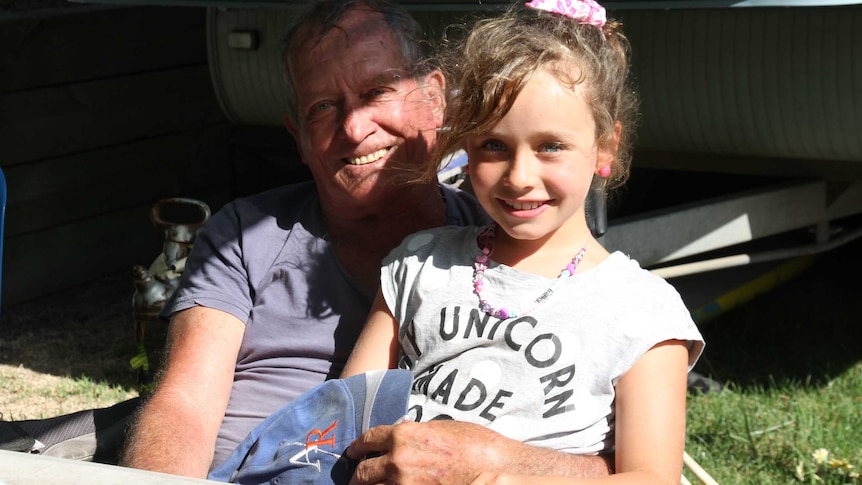 A man in his 70s sits at a caravan park with his eight-year-old granddaughter.