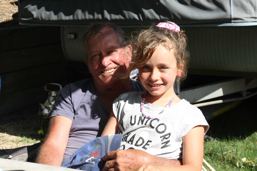 A man in his 70s sits at a caravan park with his eight-year-old granddaughter.