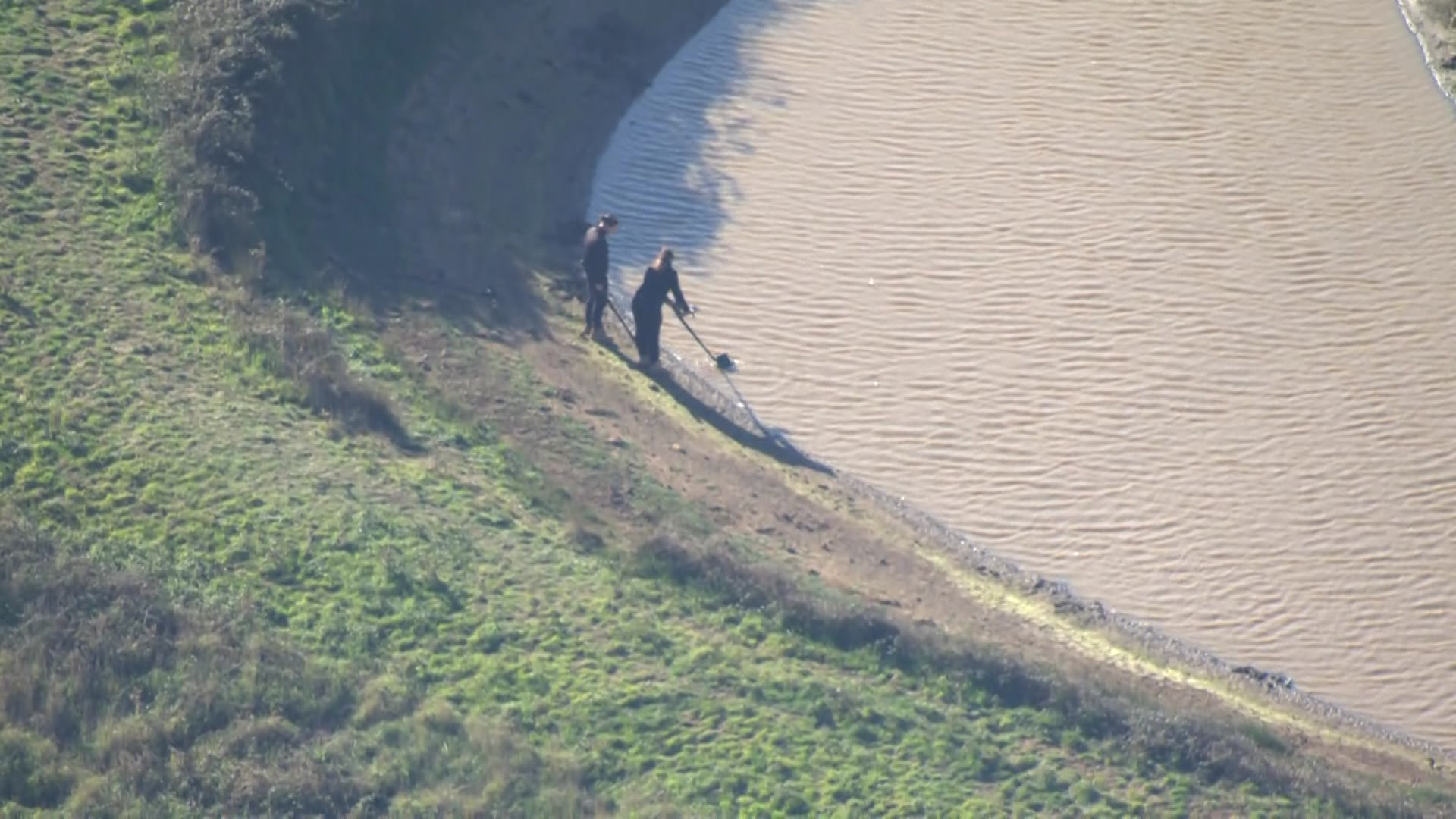 Two police officers stand by the edge of a dam, one of them holding what looks like a metal detector sifting through water