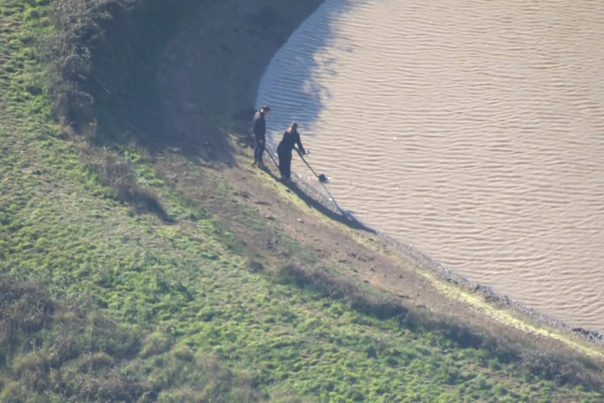 Two police officers stand by the edge of a dam, one of them holding what looks like a metal detector sifting through water
