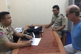 James sits handcuffed at a desk with two Cambodian policemen.