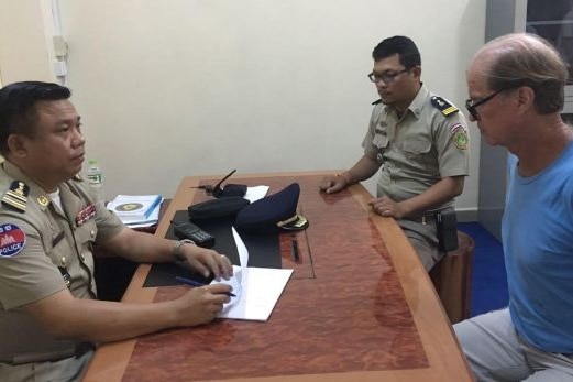 James sits handcuffed at a desk with two Cambodian policemen.