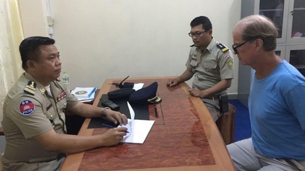 James sits handcuffed at a desk with two Cambodian policemen.