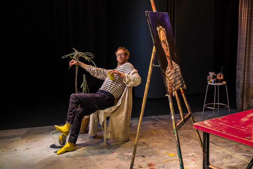 A middle-aged man with glasses leans back in a chair. Beside him is a portrait of himself, resting on a easel.