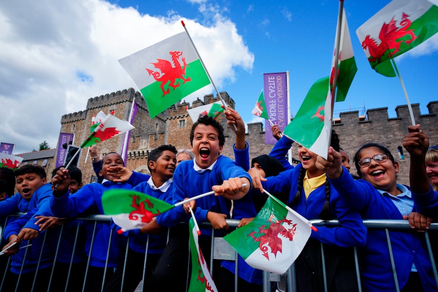 Children wave Welsh flags while standing behind metal crowd barriers outside a castle.