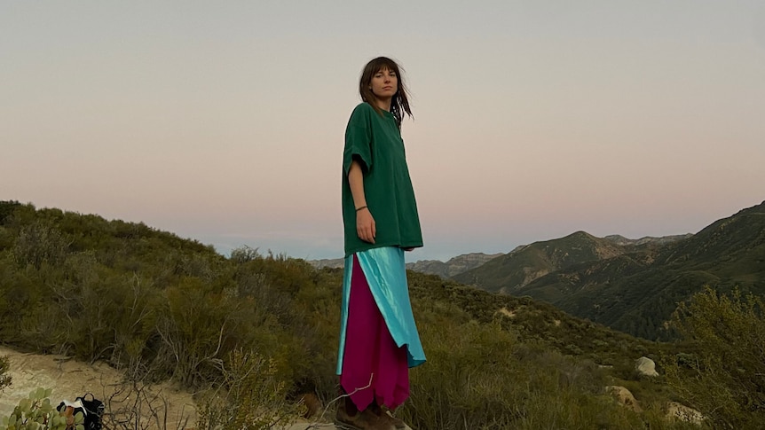 Angie McMahon stands on a hillside at dusk in an oversized green top, turquoise skirt and flowy red pants.