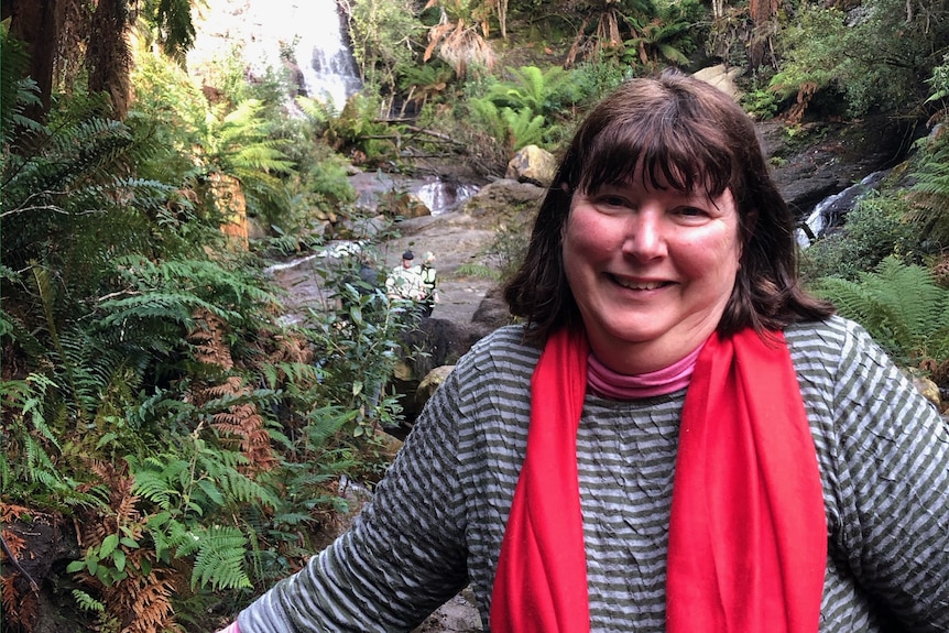 A smiling woman wearing a grey jumper and a red scarf around her neck, standing in front of ferns and a rocky hillside.