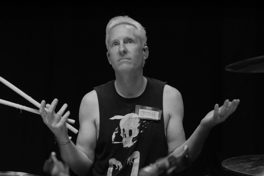 A greyscale image of Josh Freese sitting at a drum kit, shrugging wearing a "hello, my name is: Josh" sticker