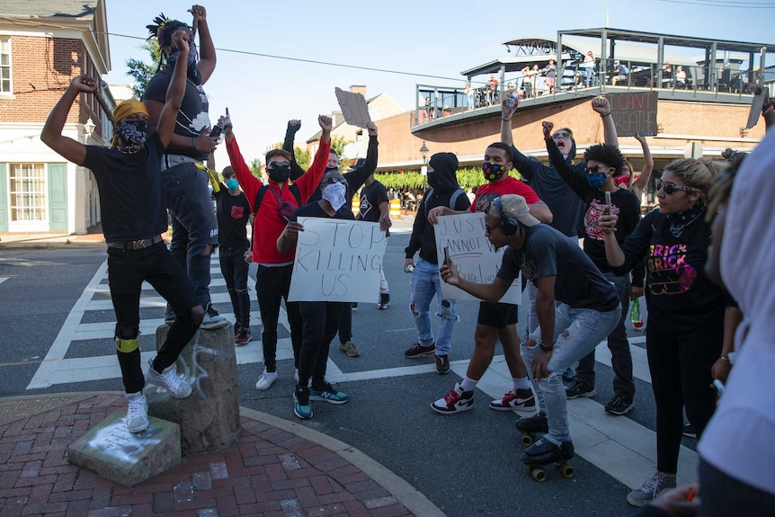 About 20 protesters stand holding placards and holding their hands in the air as they protest police brutality.