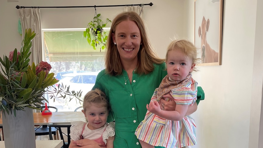 A photo of a mother in her house, holding two young female children 