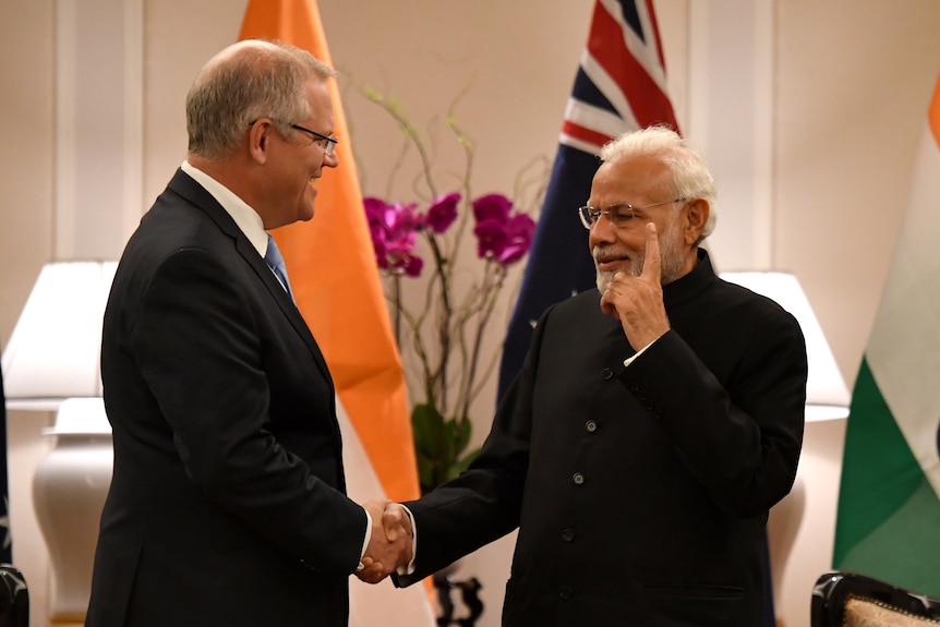 Two men wearing suits shake hands in front of the Indian and Australian flags.