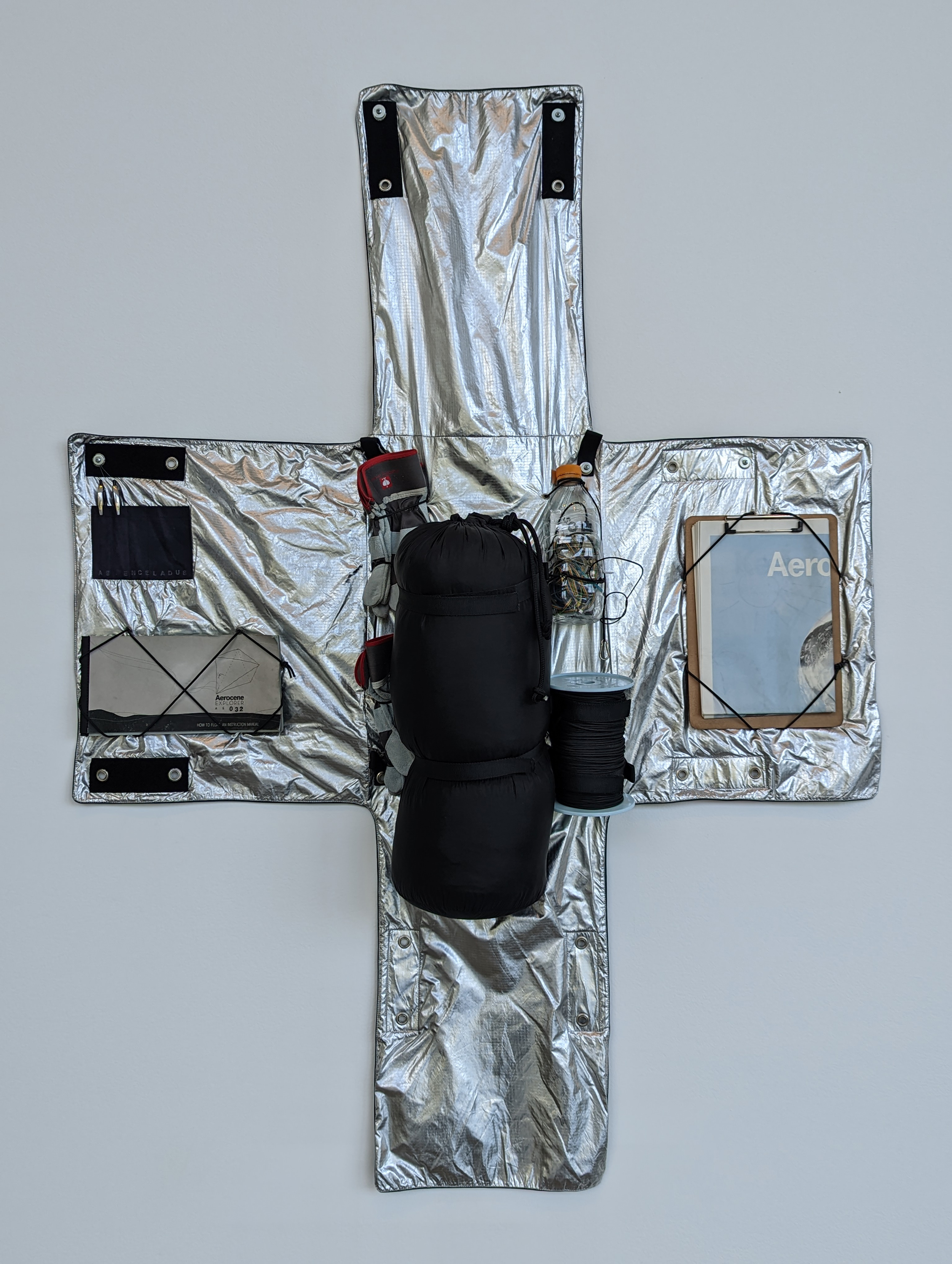 Silver fabric shaped like a cross. Inside is equipment, including a clipboard with instructions, gloves and a solar battery