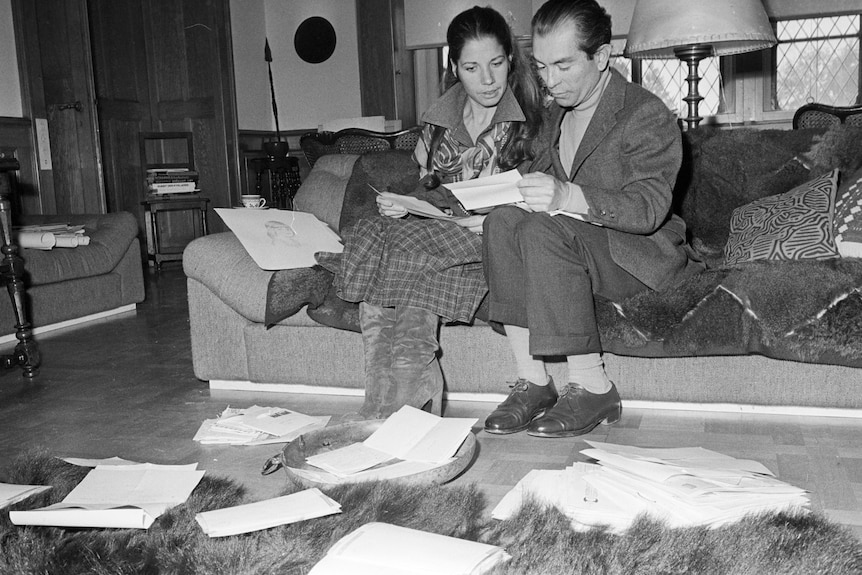 Black and white photo from 1970s of a middle aged couple sitting on a sofa surrounded by letters