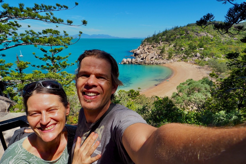 Selfie of a man and a woman smiling at the camera on a hill in front of a pristine beach