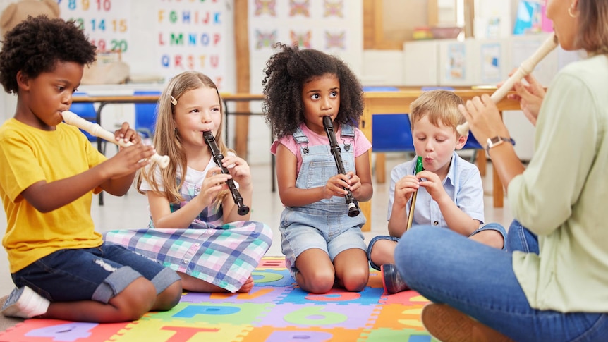 Four diverse elementary school children sit on a mat in a classroom and learn to play the recorder with a teacher.