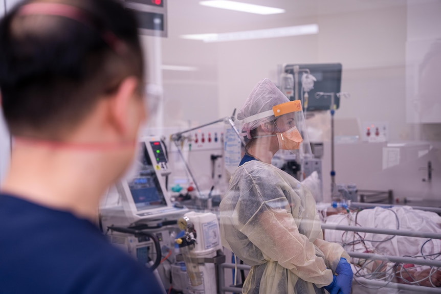 A man in hospital scrubs looks on while a women in full ppe stands.