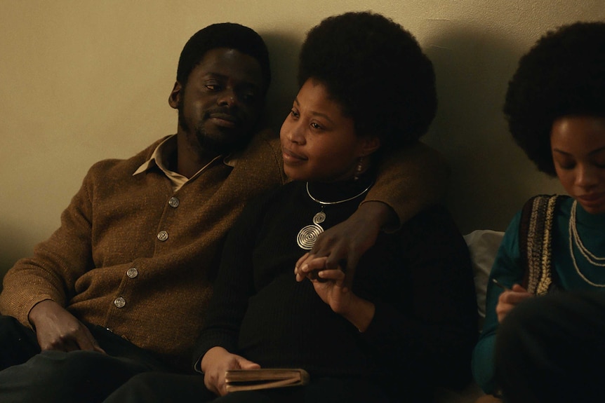 Actors Daniel Kaluuya & Dominique Fishback sitting in a loungeroom, arms around eachother, in film Judas and the Black Messiah