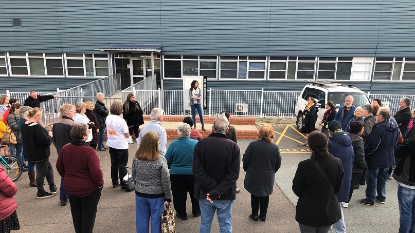 About 20 people stand in a car park, listening to a woman speak.