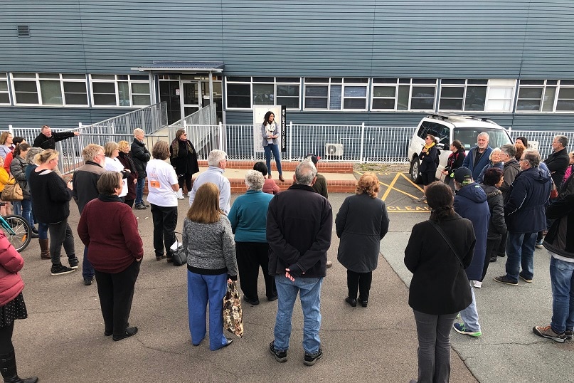 About 20 people stand in a car park, listening to a woman speak.
