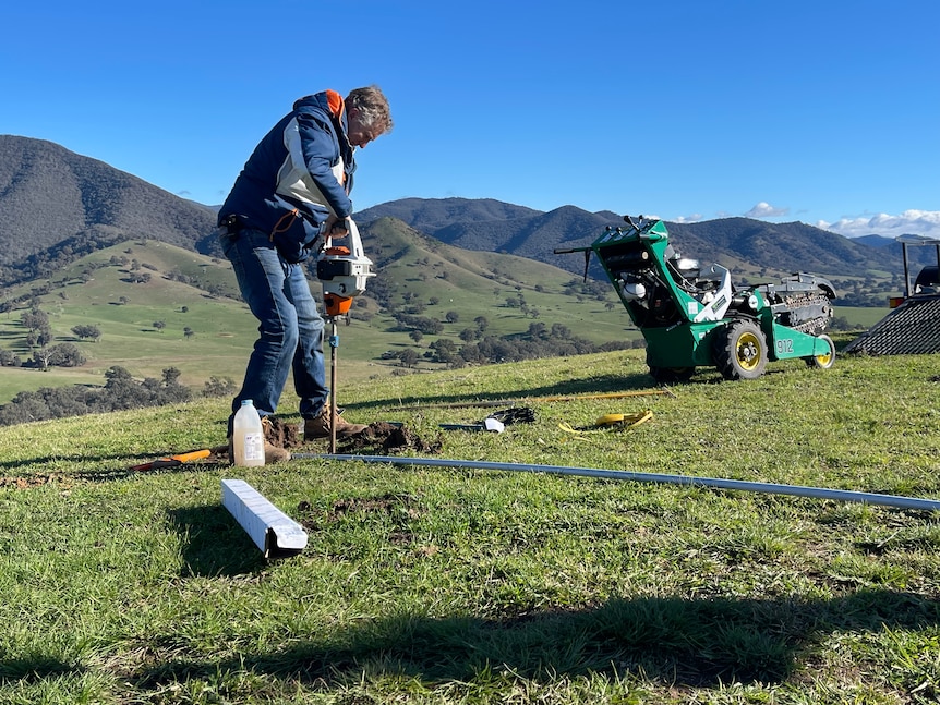A man pushes a drilling machine into the ground. He stands in a lush green paddock with a mountain range in the background.
