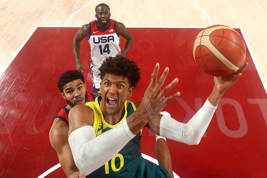 An Australian male basketballer holds the ball in his left hand as he goes up for a basket against the USA.