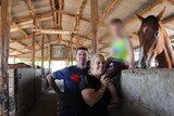 A man and woman stand next to a horse in a barn. A child is blurred in the photograph.