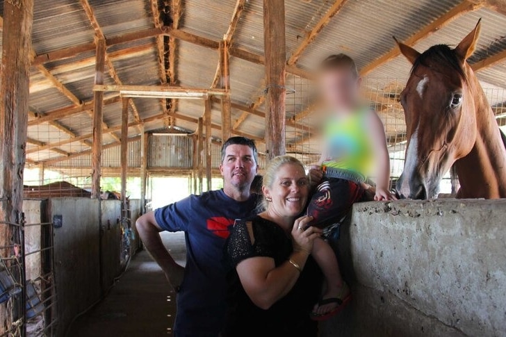 man and woman stand next to a horse in a barn. A child is blurred in the photograph.