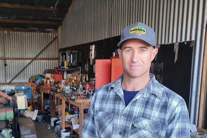 A farmer wearing a blue cap and checked shirt stands in a shed.