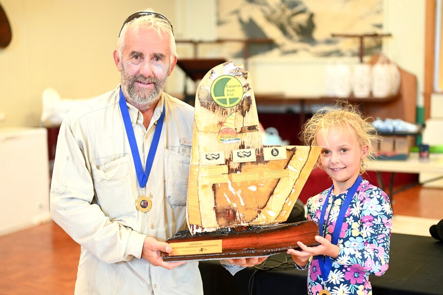A middle aged man in a fishing shirt and his young daughter in a bathing suit with gold medals hold a trophy.