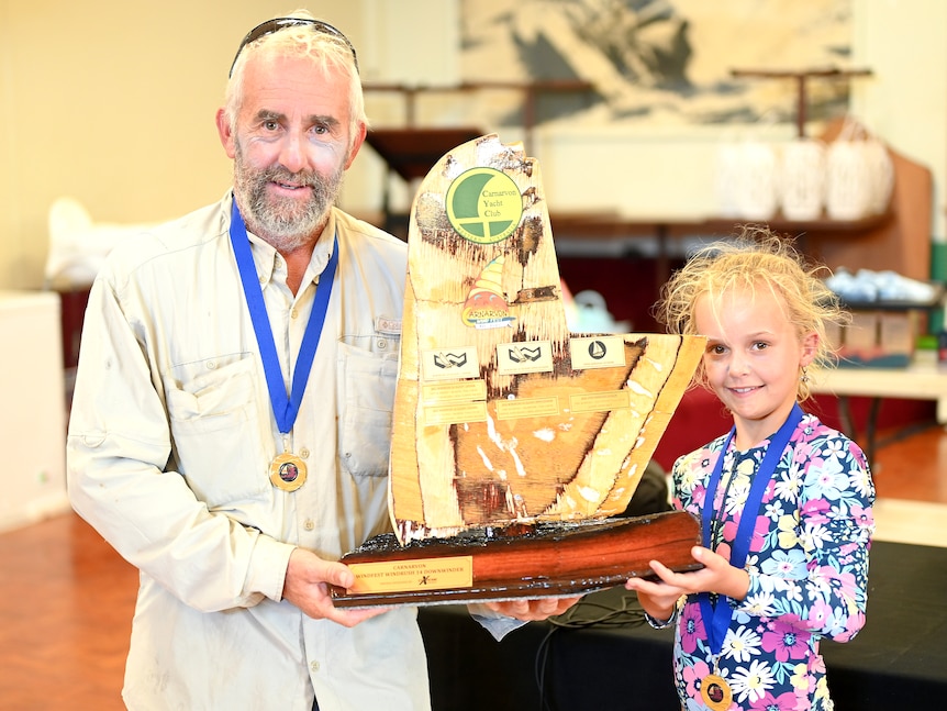 A middle aged man in a fishing shirt and his young daughter in a bathing suit with gold medals hold a trophy.