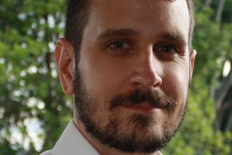 A man with a beard, moustache and short hair cut looks at the camera while standing in front of a green bush.