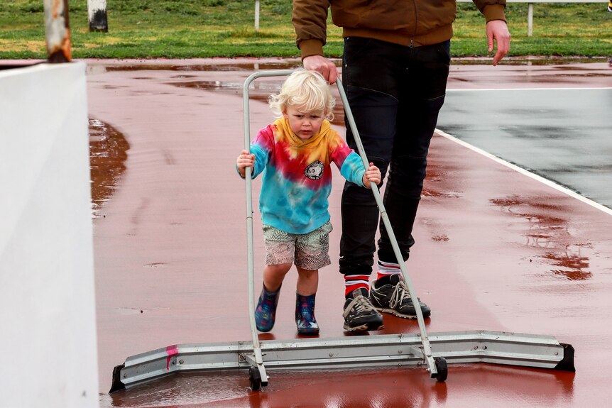 A young blonde child wearing bright coloured clothes pushes a water sweeper across a netball court