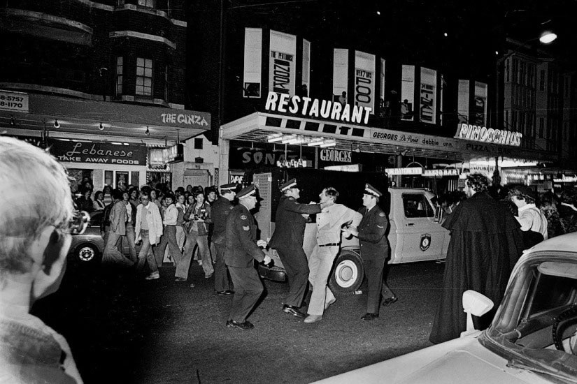 Black and white photo of police forcibly arresting a man on the street outside a restaurant.