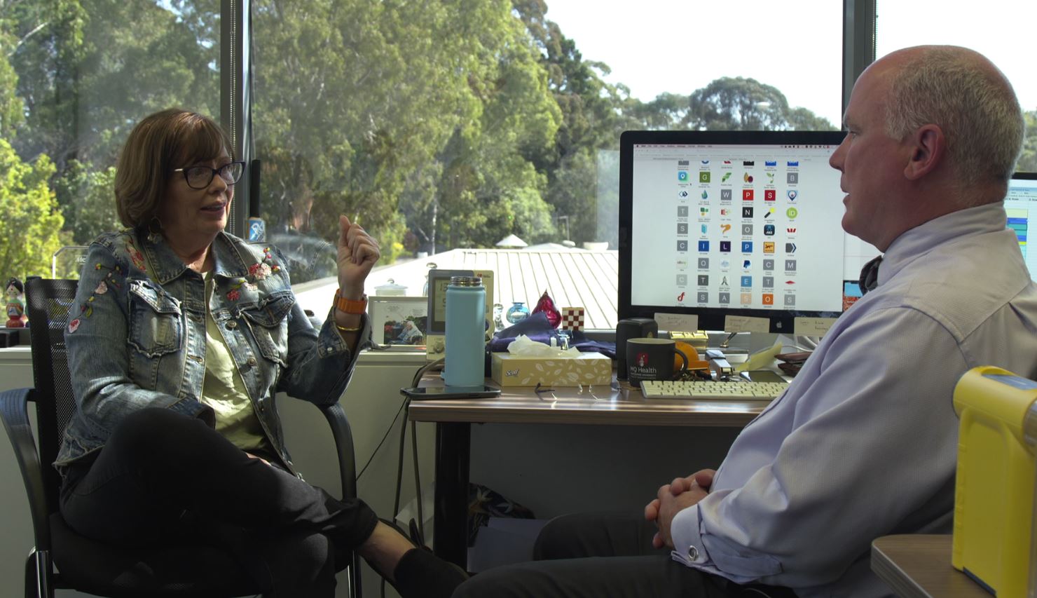 A woman and a man sitting in an office in front of a large window.