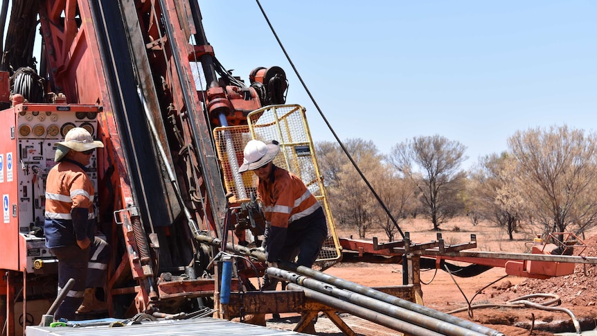 Two men dressed in orange operate a large drill rig on red dirt taking tube samples out of the machine