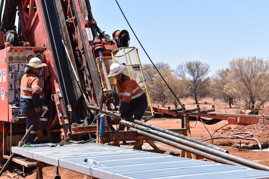 Two men dressed in orange operate a large drill rig on red dirt taking tube samples out of the machine