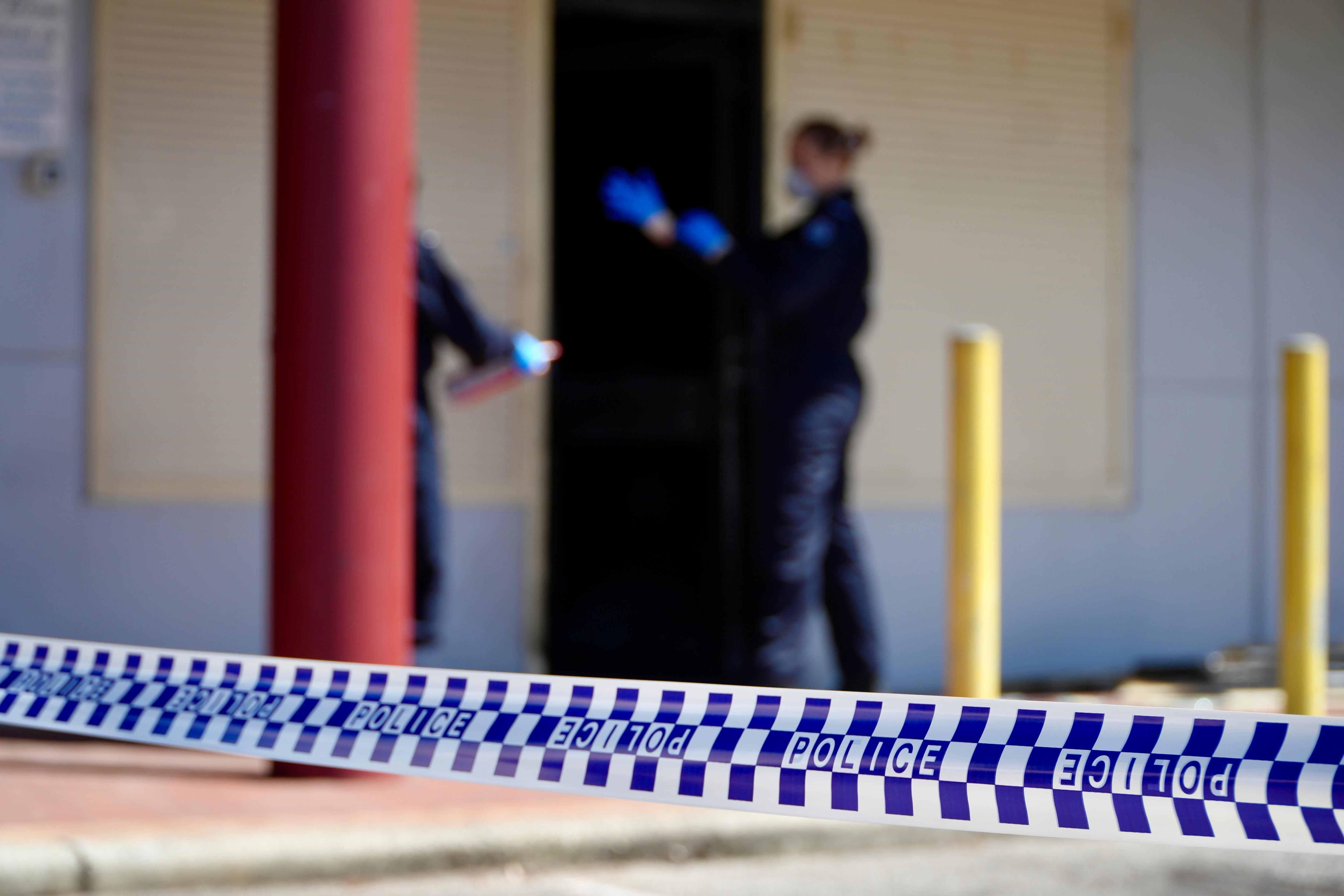 Una cinta policial está enfocada en primer plano, con el exterior de una tienda quemada al fondo con agentes de policía con trajes forenses azul marino.