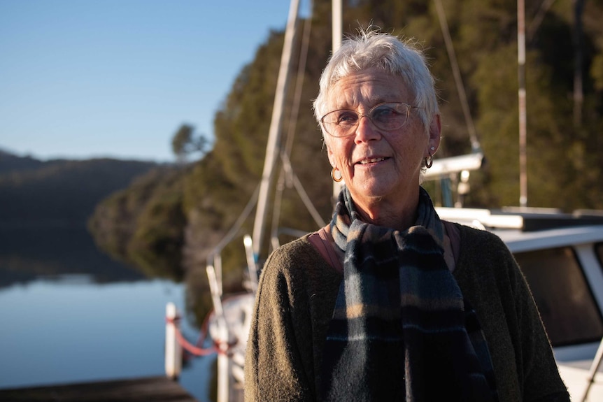 A woman wearing glasses, white hair, scarf and dark green jumper stares into the distance, a river and yacht are behind her.
