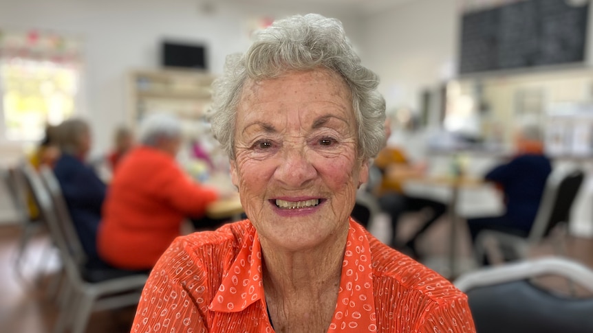 An elderly woman wearing an orange sweater smiles for the camera