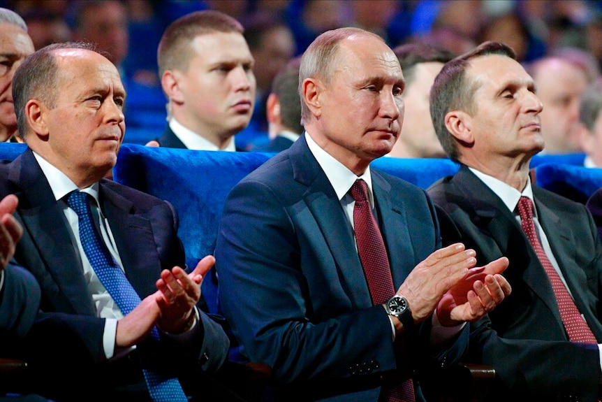 Vladimir Putin claps his hands while seated in an electric blue auditorium surrounded by other men in suits.