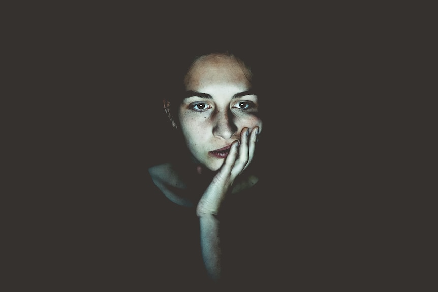 In a dark room, a woman stares into the glow of a laptop screen, looking serious, with her hand held to her mouth.