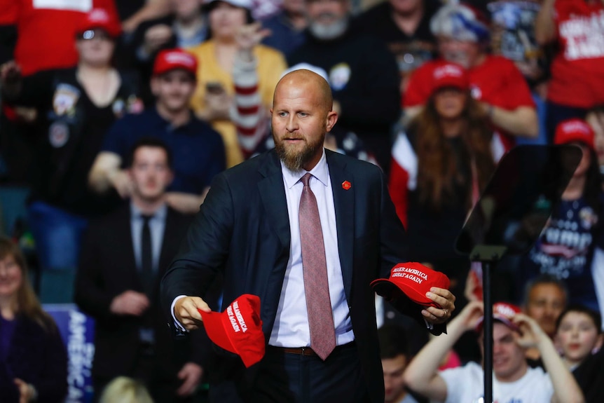 A man in a suit hands out red baseball caps while on a stage.