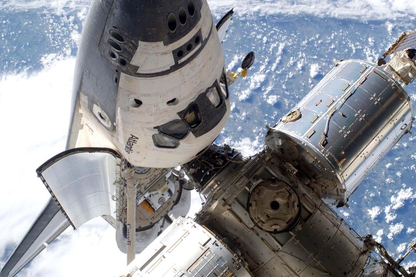 A space shuttle docks with the International Space Station, with a view of an ocean on Earth below.