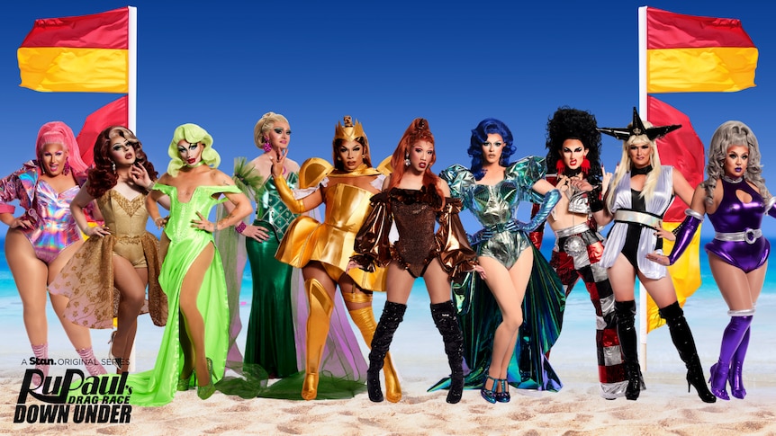 A group of 10 drag queens in various gowns, leotards and outfits pose against a beach backdrop