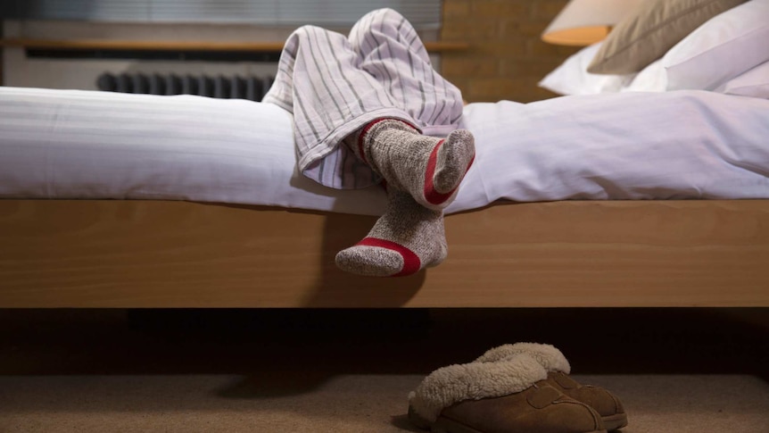 A person hanging their legs off the side of a bed wearing pyjamas and socks with slippers on the floor below
