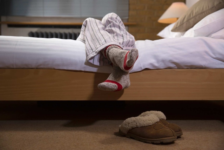 A person hanging their legs off the side of a bed wearing pyjamas and socks with slippers on the floor below