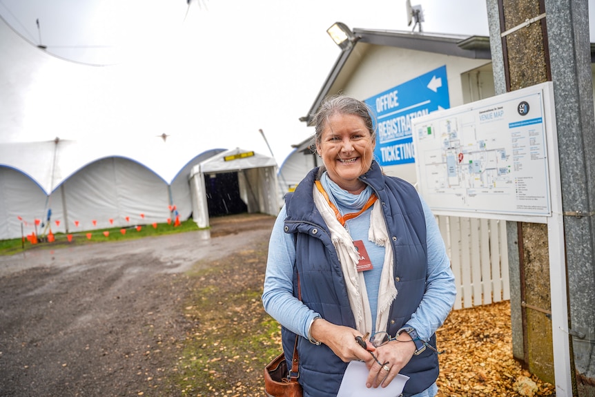 A smiling, middle-aged woman with grey hair, dressed for warmth, stands outside a giant tent.