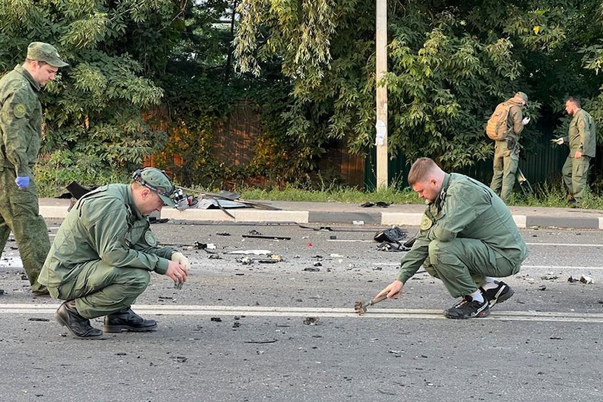 Two men dressed in army uniforms crouch on the road looking at debris. 