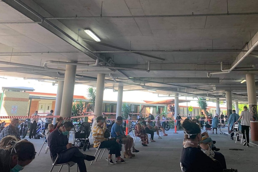 People in masks sitting on chairs spaced across an sub-floor driveway at a hospital