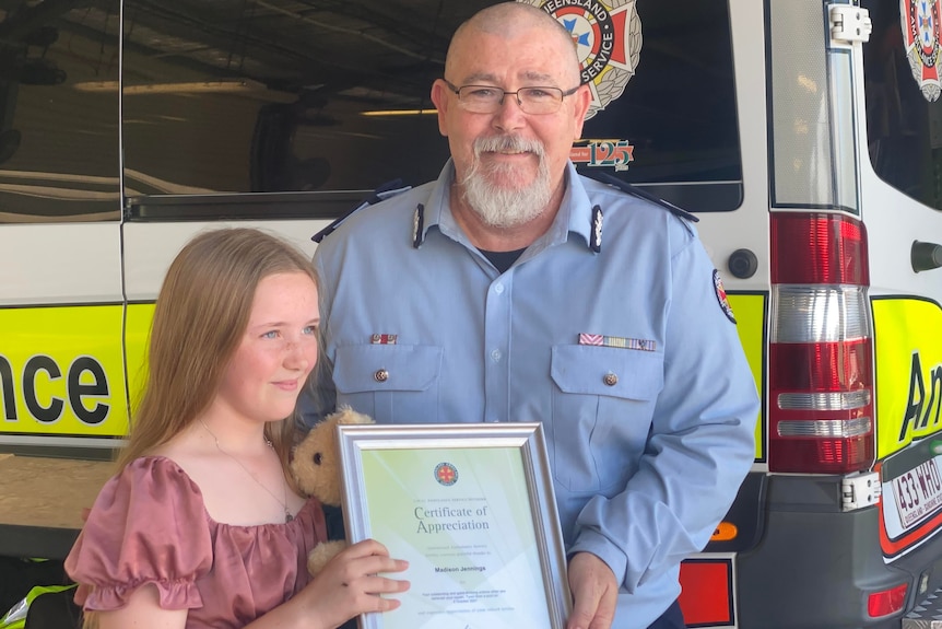 A young girl and an old man stand in front of an ambulance holding a certificate